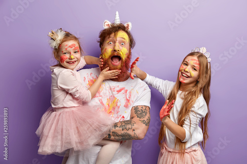 Emotional bearded busy father spends time with two naughty daughters who leave palm prints on his beard and clothes, learn how to paint, dressed in festive clothes, stand indoor. So colourful!