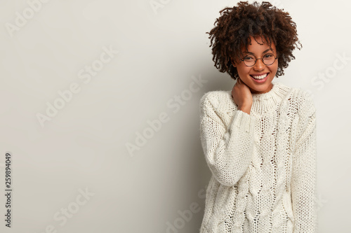 Glad laughing curly woman giggles as heard funny joke, wears white sweater, keeps hand on neck, expresses positive emotions, stands indoor, blank space right for your advertisement. Happiness
