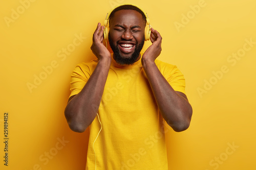 Monochrome shot of overjoyed pleased Afro American man enjoys perfect loud sound in new headphones, dressed in yellow t shirt, has free time, entertains himeslf with music. Happy expression.