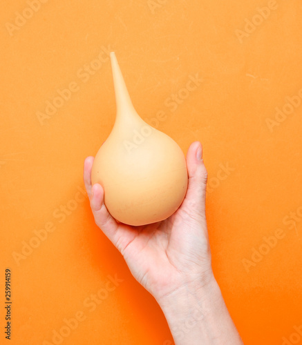 Female hand holds orange medical enema on orange background. Top view, minimalism.