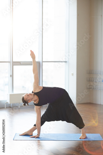 Serious handsome young man with ponytail standing on yoga mat and keeping arms outstretched while performing extended triangle pose