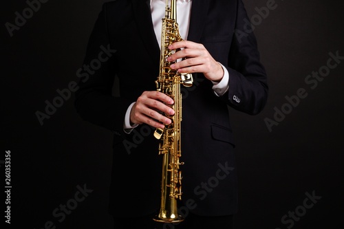 Male saxophonist in a black classic suit playing on the soprano saxophone while standing on a black background