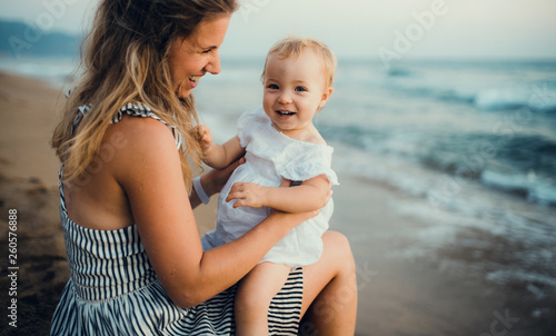 Young mother with a toddler girl sitting on beach on summer holiday.