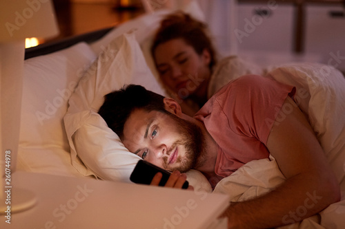 technology, internet addiction and cheat concept - man using smartphone at night while girlfriend is sleeping