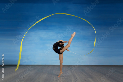 Girl gymnast in a black gymnastic swimsuit makes a circle in the air with a yellow ribbon on a blue sky background.