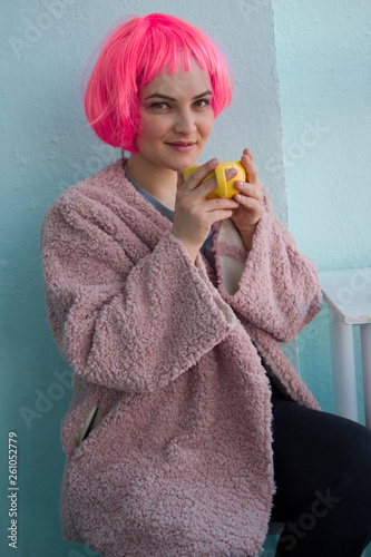 beautiful young woman with pink hair in pink fur coat sitting in coffee shop and holding yellow coffee mug