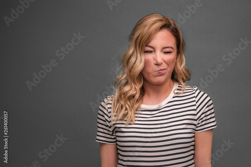 A young woman is squinting a sour grimace while standing against grey background.