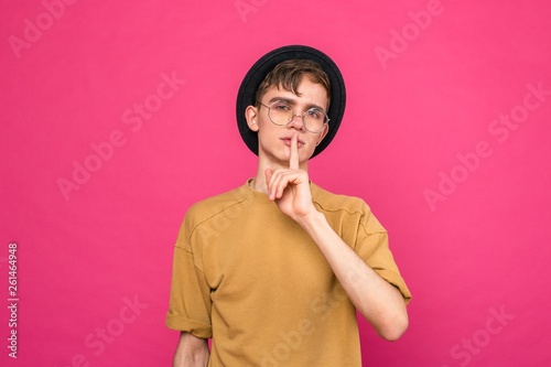 Portrait of a young man in a black hat pointing the silence with a finger to his lips standing on a pink background