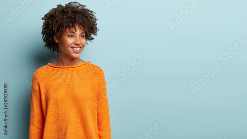 Image of dreamy woman with crisp hair looks pensively aside, has gentle smile on face, wears orange jumper, imagines something pleasant, models over blue background, blank space for promotion