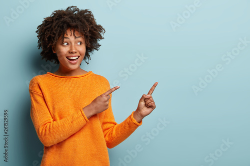 Horizontal shot pleased dark skinned female with Afro haircut, points away with both fore fingers, shows blank space for your promotion, isolated over blue background. People, advertisement concept