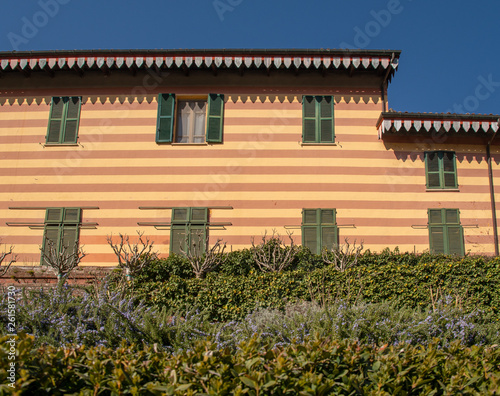 Close-up of an old building with a striped façade, green wooden shutters and hedges in the foreground in springtime, Piedmont, Italy