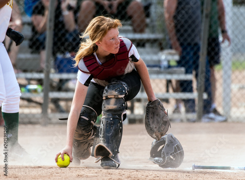 Skilled softbaall catcher with red hair and protective gear gaining a grip on the loose ball while looking up field.