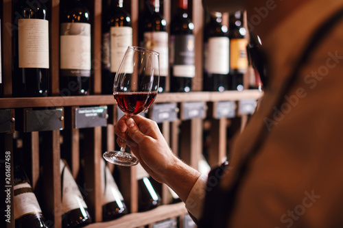 Bartender or male cavist standing near the shelves of wine bottles holds a glass of wine, looks at tint and smells flavor of wine in glass.