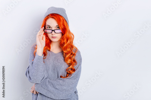 Thinking serious skeptical redhead teen girl wearing glasses dressed in knitted sweater and beanie hat on gray background