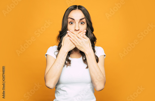 Oops... Shocked girl with big dark brown eyes near the flame-colored background clothed in white T-shirt is holding her hand over her mouth because of shock.