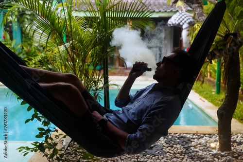 Man resting in a hammock and vaping and letting off steam from an electronic cigarette