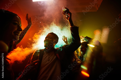 Crowd of cheerful hilarious young multiethnic people drinking alcohol and dancing in smoke at noisy party