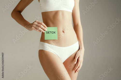 Young adult woman holding card with word DETOX. Woman health. Female model holding card near stomach isolated on gray studio background. Cut out part of body. Medical problem and solution.