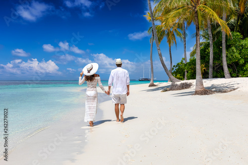 Elegant traveler couple walks down a tropical beach with coconut palm trees and turquoise waters in the Maldives islands