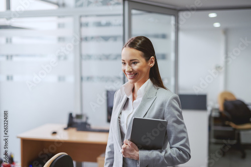Beautiful Caucasian brunette in formal wear standing in office with tablet in hands. Great things never came from comfort zone.