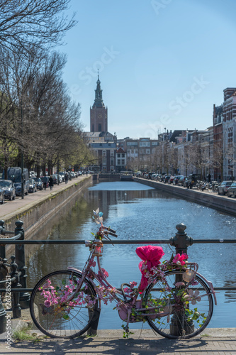 Pink and decorated bicycle lays on the bridge over a calm canal against a lamp post reflected on the water with the ST James church in the background, The Hague, Netherlands
