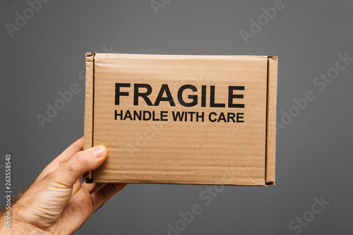Fragile handle with care warning text on cardboard with text sign in man's hand against gray background - transportation of delicate objects and good