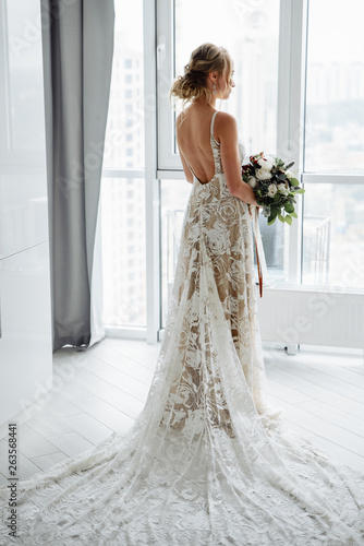 Gorgeous bride in a luxurious long dress with a bouquet in hand, stands and looks out the window