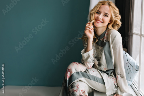 Beautiful young woman with long blonde wavy hair sitting on window sill in room with turquoise wall. She's smiling and enjoying morning time. Wearing nice pajama.