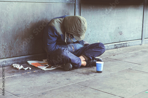 Homeless adult man sitting on the street in the shadow of the building and begging for help and money. Problems of big modern cities. Indifference of people. Social issues.