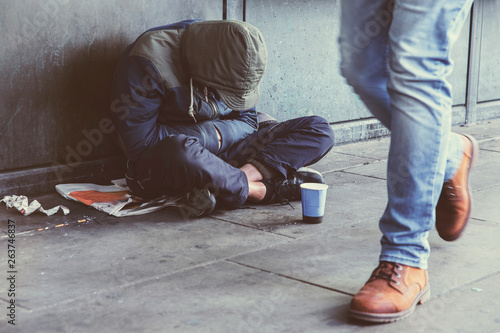 Homeless adult man sitting on the street in the shadow of the building and begging for help and money. Problems of big modern cities. Indifference of people. Social issues.