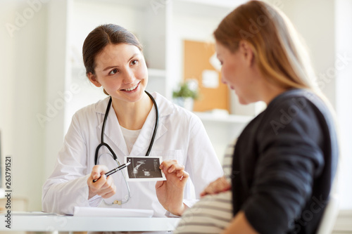 Portrait of smiling onstetrician showing ultrasound scan of baby to pregnant woman visiting doctor, copy space