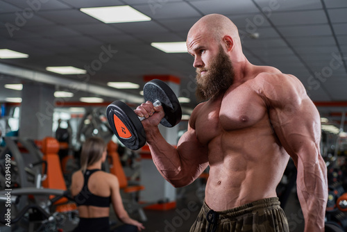 Brutal, muscular, bald with a beard athlete in the gym. Lifts heavy weights, trains the biceps. Beautiful chest muscles