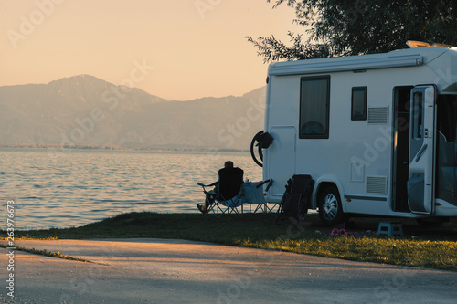 Man sitting near his caravan observes the sunset close the lake