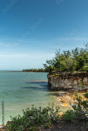 Darwin Australia - February 22, 2019: Detail of East Point Shoreline shows brown, yellow and darker rocky cliffs covered by green vegetation under blue sky and greenish sea water of Beagle Gulf.