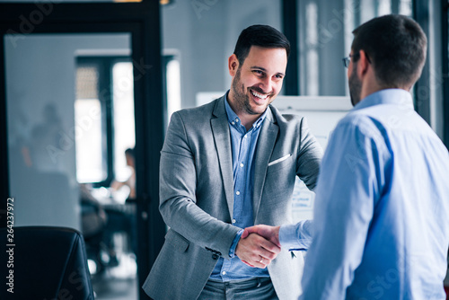 Two smiling businessmen shaking hands while standing in an office.