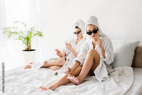 stylish women in bathrobes and sunglasses, towels and jewelry lying in bed, doing pedicure, smoking and using smartphone