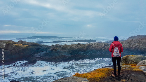 A girl wearing pink jacket and a backpack, standing at the stony shore and looks at the sea. Rough surface of the sea, waves struggle through the rocks to get to the shore. Solo traveling girl.