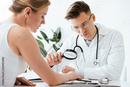 selective focus of handsome dermatologist in glasses holding magnifying glass while examining woman in clinic