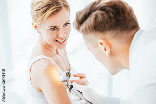 selective focus of cheerful woman looking at dermatologist holding dermatoscope while examining hand