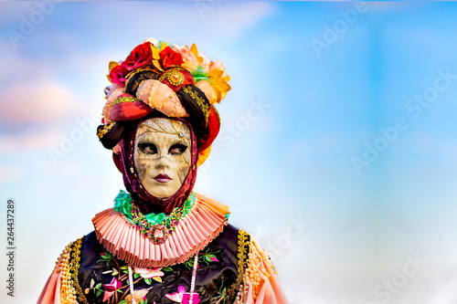A young girl with a curious look in a pink flower suit and a headdress with colorful flowers, and a mask at the carnival in Venice