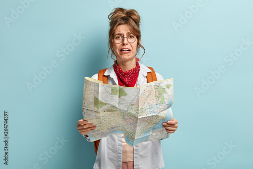 Crying female tourist lost in unknown destination, holds map, tries to find way, looks with dissatisfied dejected expression, wears round spectacles, has rucksack with packed things, stands indoor