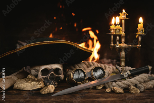 Pirate table with treasure gold, sword, binoculars, human skull, captain hat and scroll map on a burning fire background. Piracy concept.