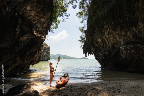 Blonde Caucasian woman with a paddle standing next to sea kayak at secluded beach in Krabi, Thailand