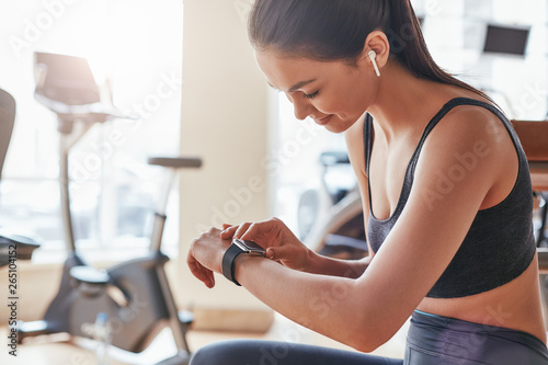 Checking results. Side view of beautiful young woman looking at her sport bracelet while having a rest at gym