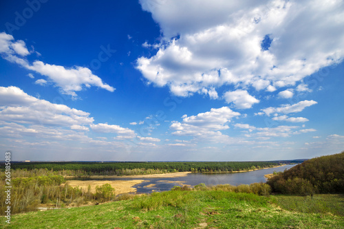 Beautiful view of river, green forest and blue cloudy sky from the top of the hill. Spring landscape.