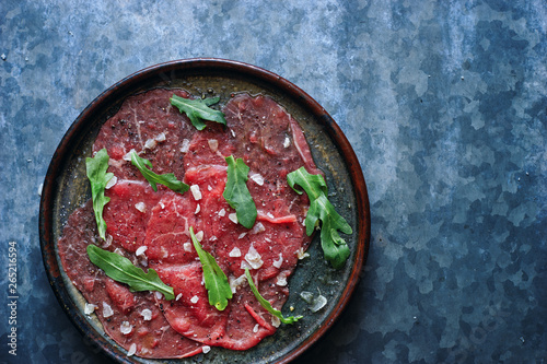 Raw marbled beef carpaccio with parmesan cheese, different spices and arugula in a plate top view space for text