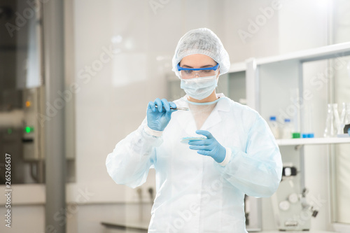 Serious concentrated young female laboratory scientist in disposable cap and coat using tweezers while taking medical sample from petri dish