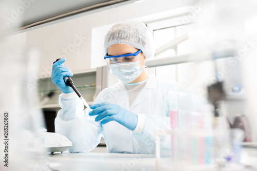Serious concentrated female microbiologist in sterile clothing and safety goggles sitting at table and dropping reagent in petri dish while doing research in laboratory