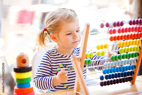 Adorable cute beautiful little toddler girl playing with educational wooden rainbow toy pyramid and counter abacus. Healthy happy baby learning to count and colors, indoors on sunny day.