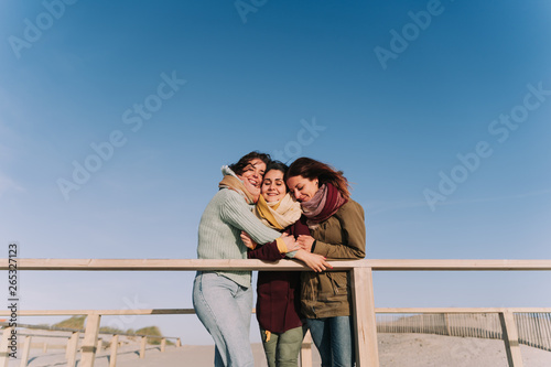 Three friends hugging each other in a spring-like afternoon. Two of them are wearing dark clothes and the other more light colors. They look happy and comfortable together